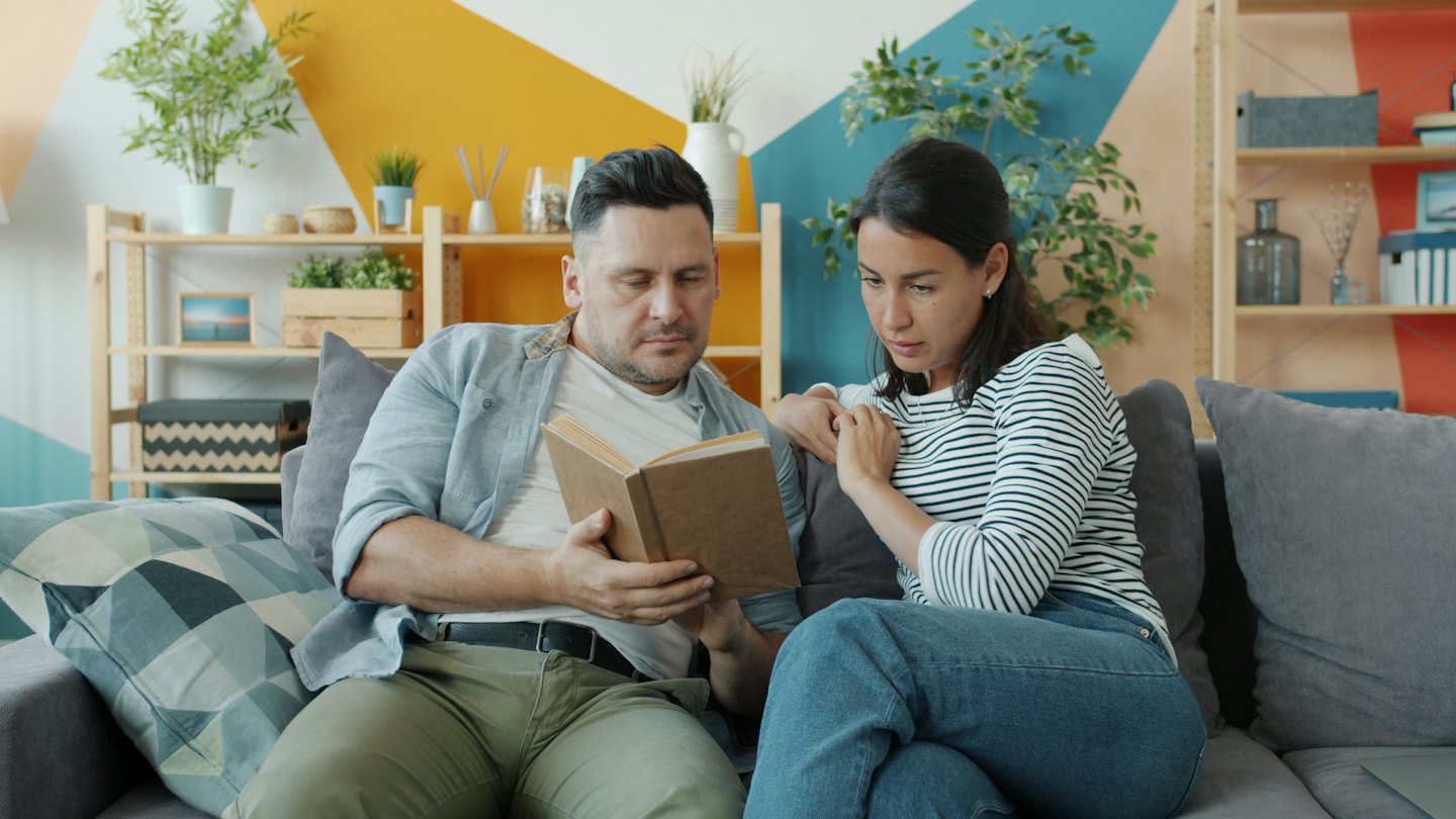Ein Paar sitzt auf einem Sofa und liest gemeinsam ein braunes Buch.